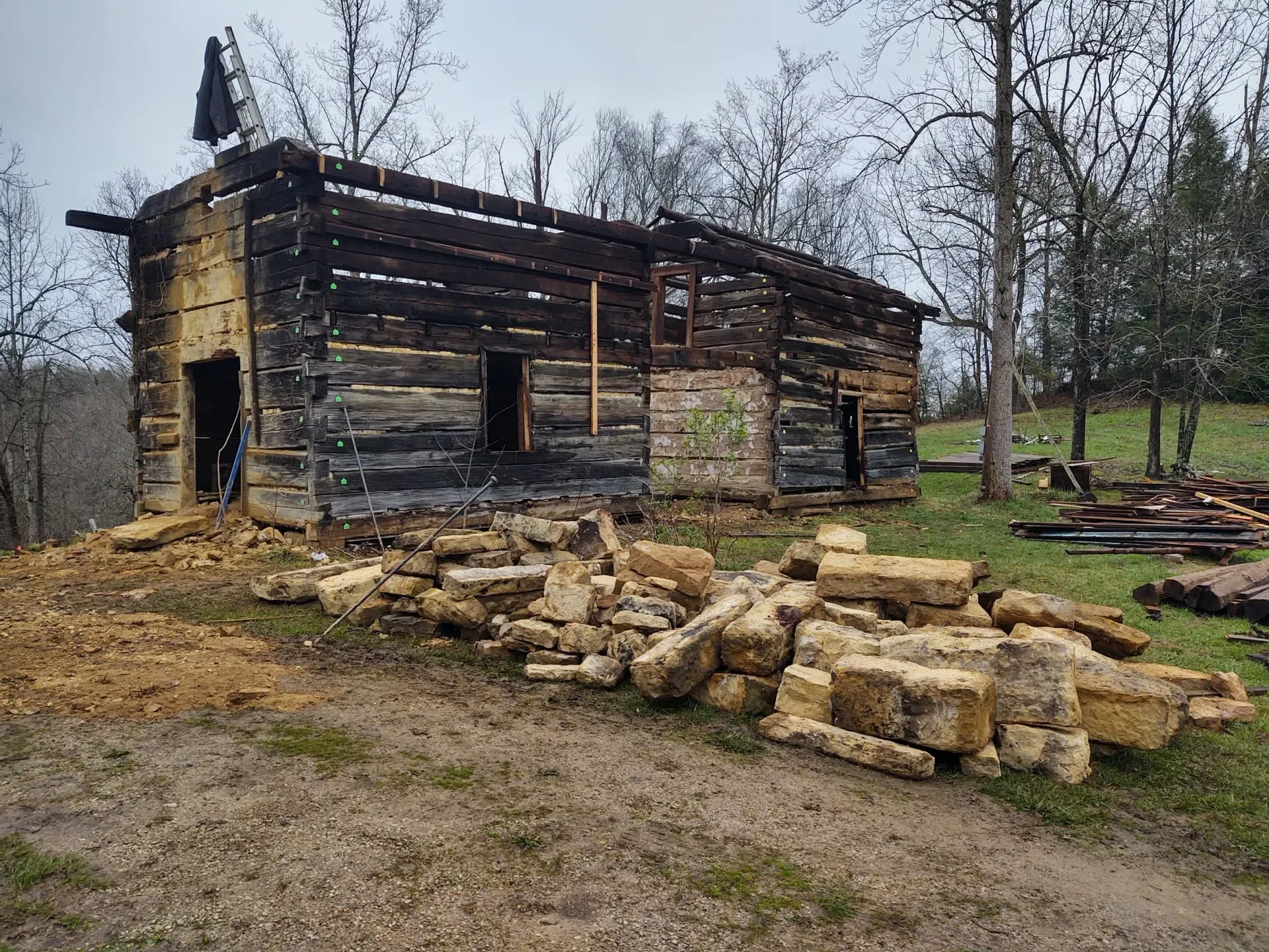 Charred wooden cabin ruins with scattered logs in a muddy yard under bare trees