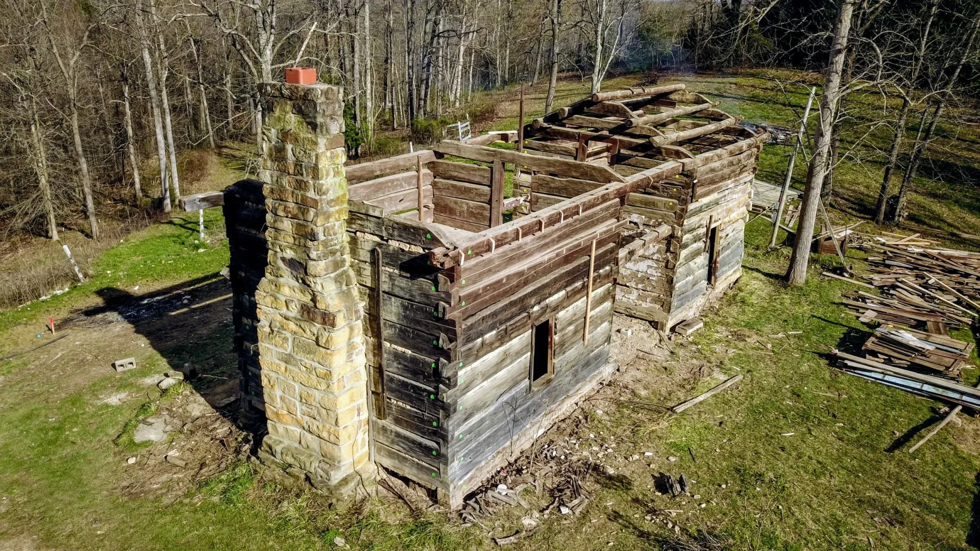 Aerial view of a small abandoned wooden building with a chimney, surrounded by trees and grass.