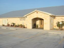 Stone building entrance with arched doorway and small sign, viewed from a parking lot.