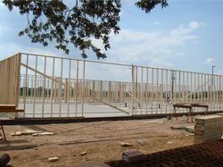 Empty fenced construction area with a concrete slab, white metal frame, and dirt foreground under a tree.