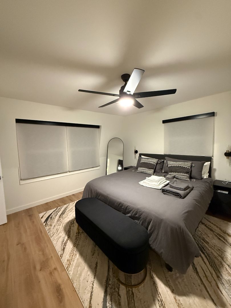 Modern bedroom with a gray bed, ceiling fan, bench, rug, and two windows with blinds