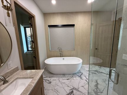 Black roller shade covers a bathroom window above a bathtub. White walls and marble tile are visible.