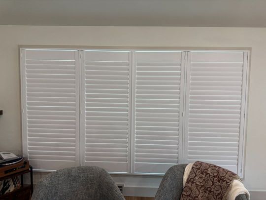 White plantation shutters covering a large window behind two chairs in a living room