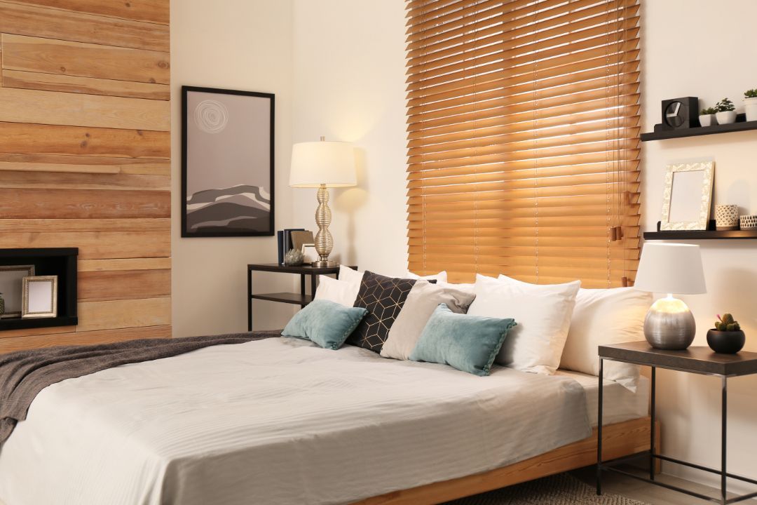 Cozy bedroom with a white bed, teal pillows, wood accent wall, lamp, and woven blinds by a window