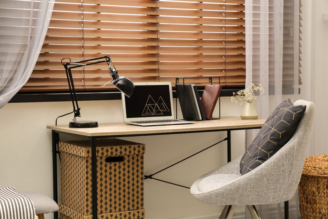 Cozy home office with a desk lamp, books, and a gray chair by a window with blinds