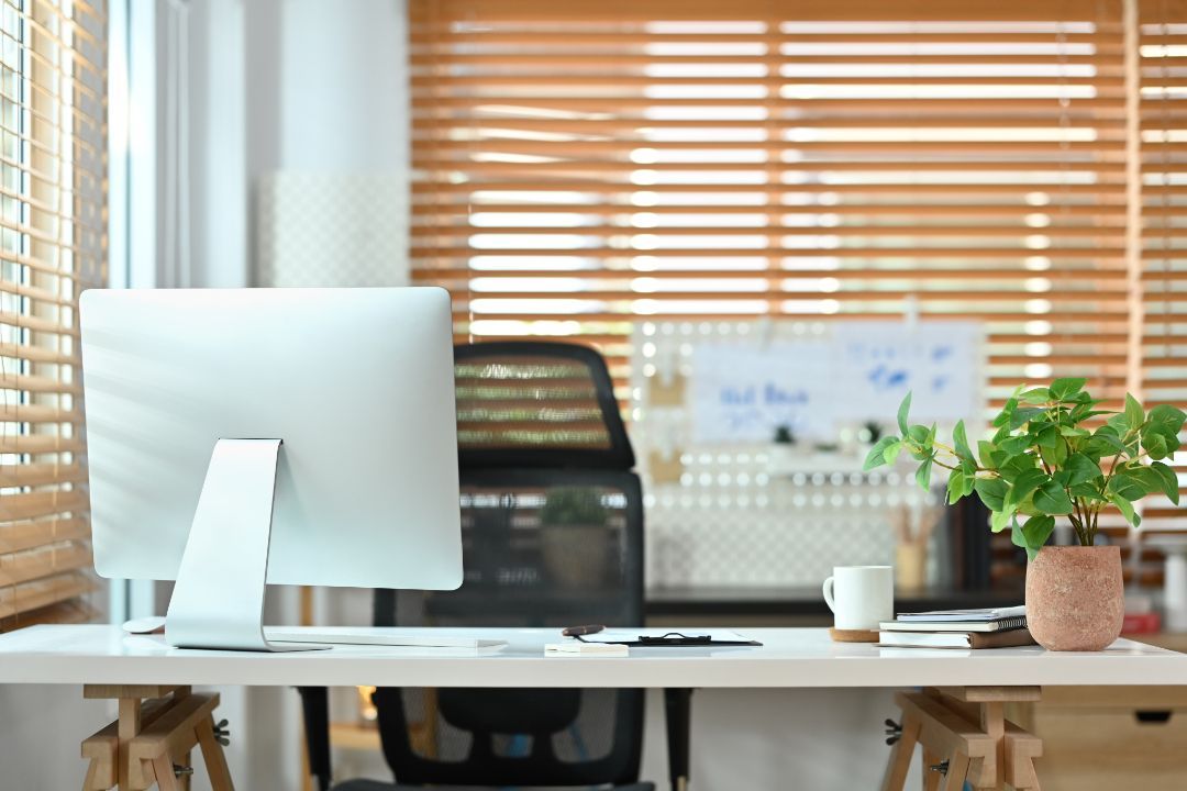 Bright office desk with computer, chair, coffee cup, and potted plant beside window blinds.