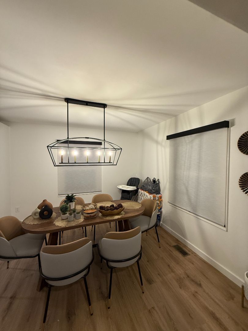 Dining room with round wooden table, four chairs, black pendant light, and a window with blinds.