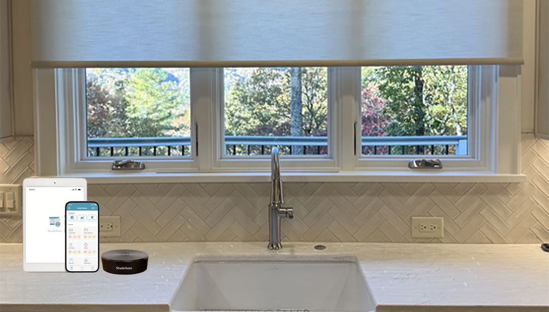 Kitchen sink beneath a wide window with white blinds, showing a smartphone and smart speaker on the left.