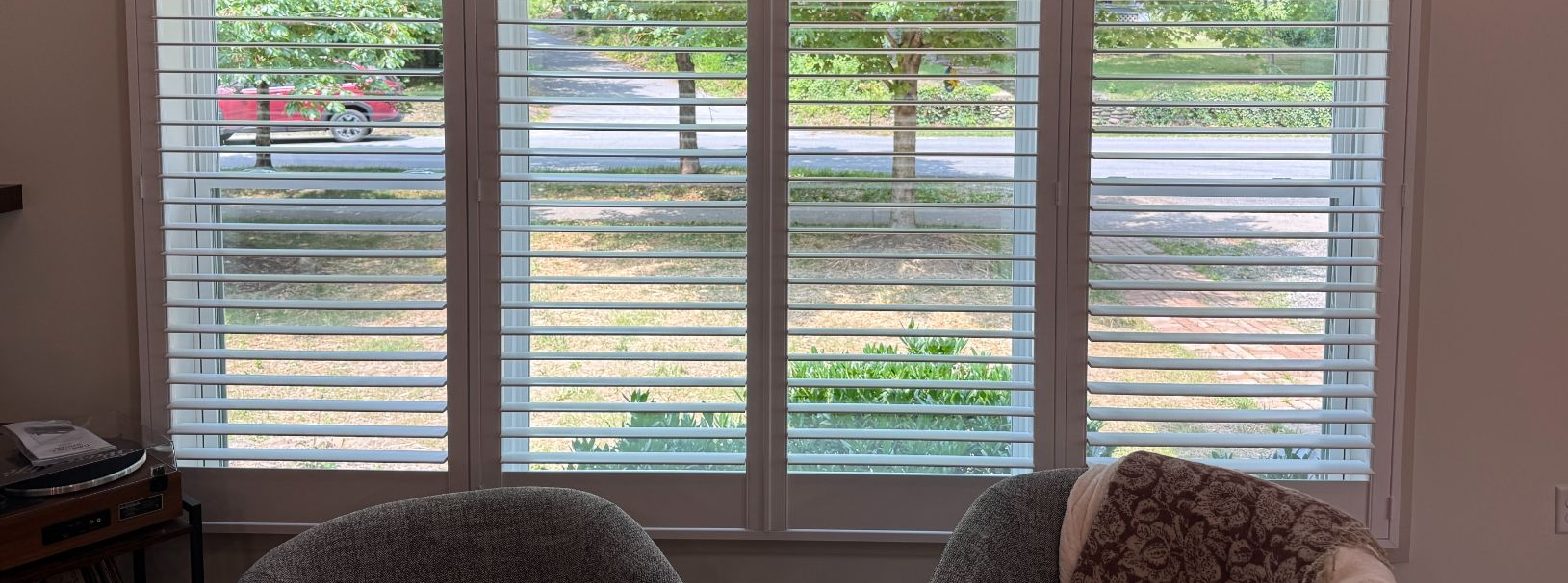 Living room window with white shutters, overlooking a street and greenery