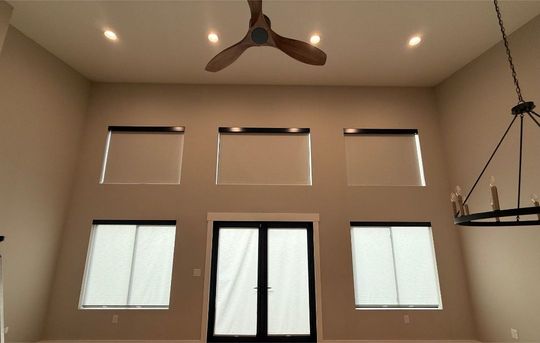 Bright beige room with tall windows, double doors, ceiling fan, and hanging chandelier.