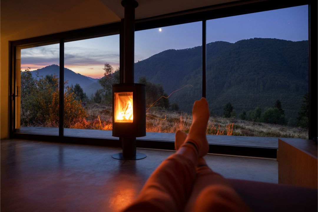 Cozy cabin view with a glowing lantern, mountains at dusk, and a person’s feet by the window