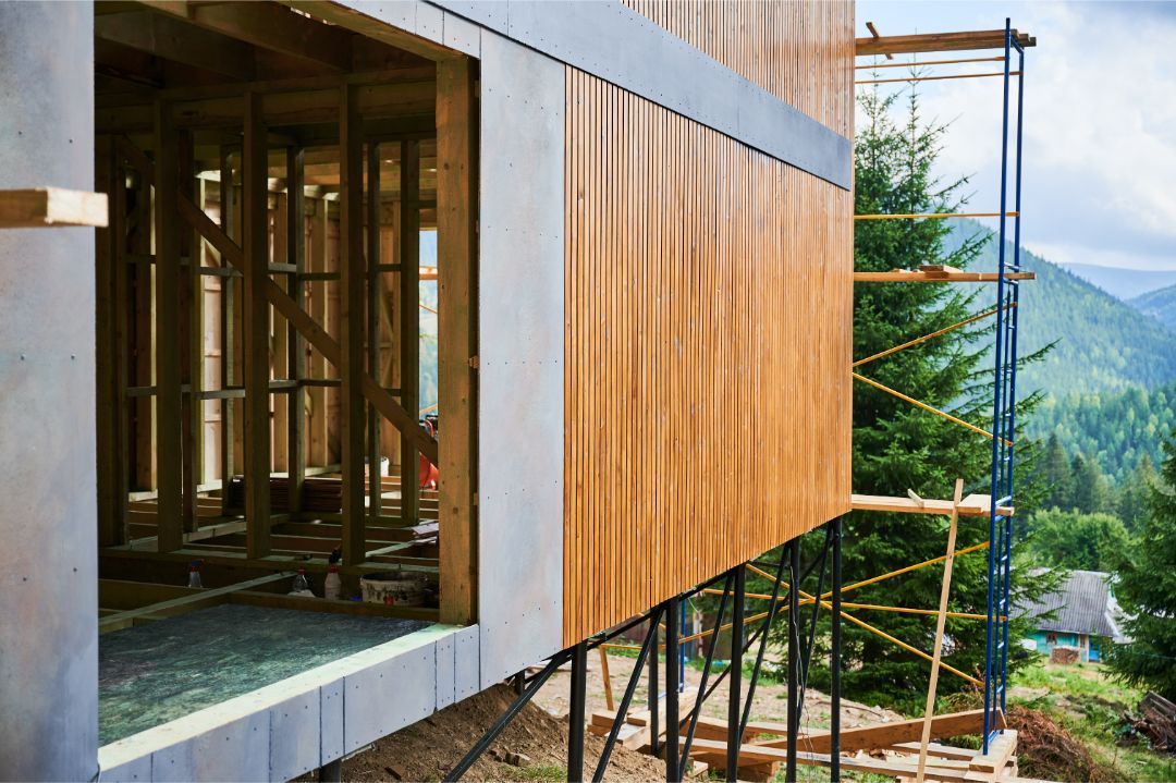 Modern house exterior with wood siding, large window, and mountain view beyond the scaffolding