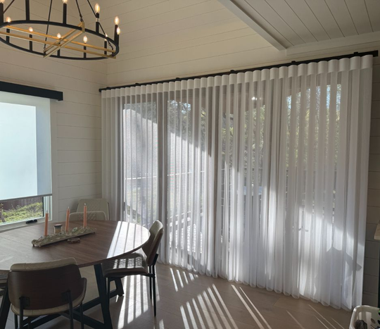 Dining room with round table, chandelier, and white vertical blinds casting sunlight and shadows across the floor