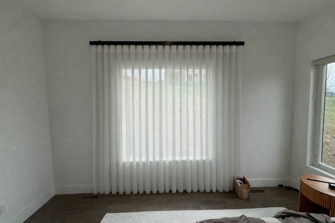 Empty white bedroom with a centered window, sheer curtains, and a small wooden side table.