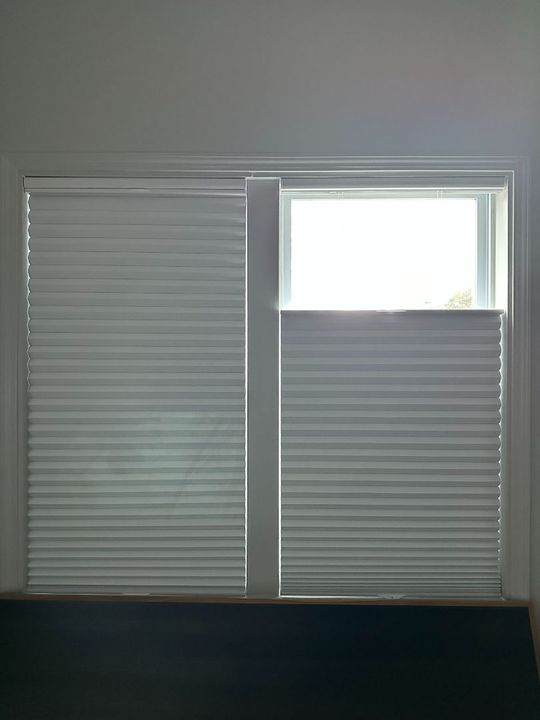 Closed white window blinds covering a wall, with a small open upper window on the right
