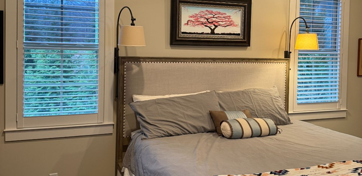 Cozy bedroom with a gray bed, striped pillows, framed art, and two windows with custom interior shutters.