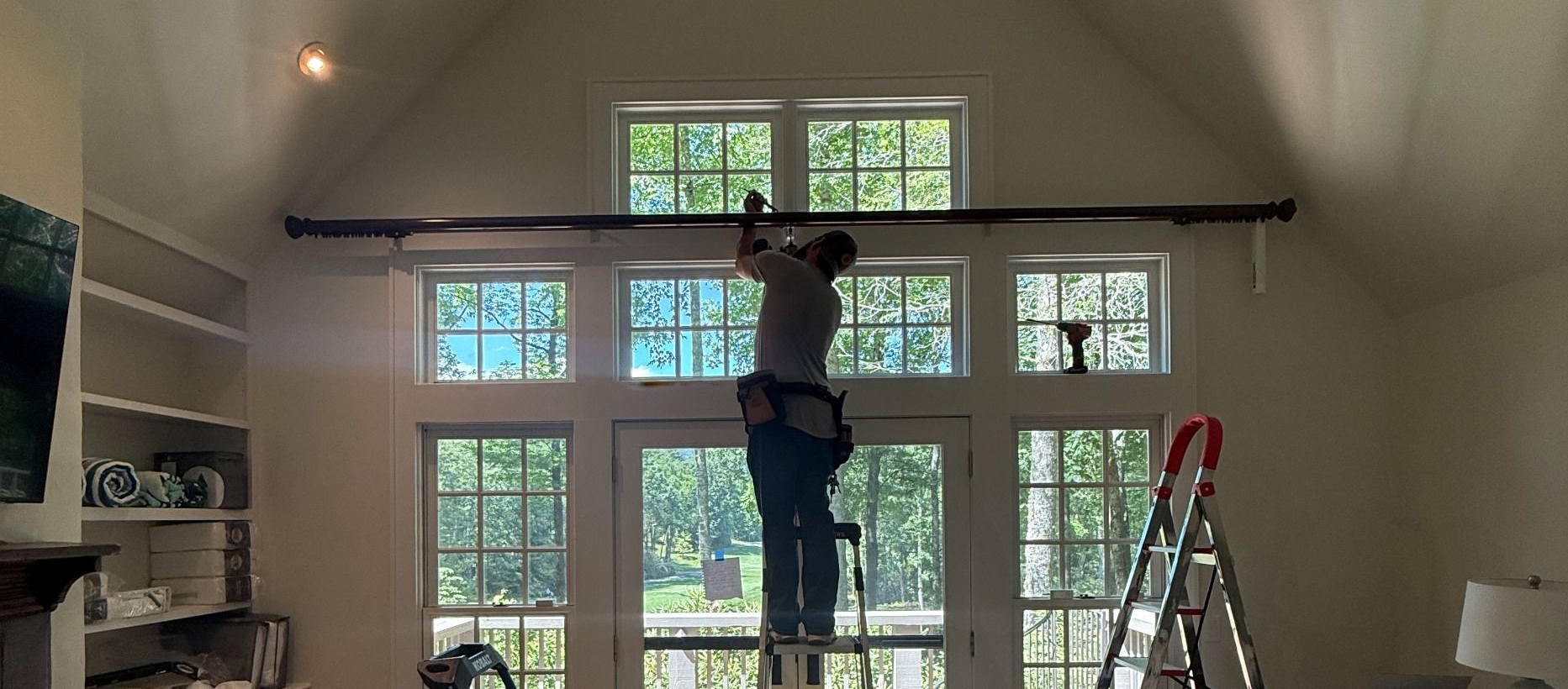 AVL Installer on a ladder installing window treatments above tall windows in a bright room