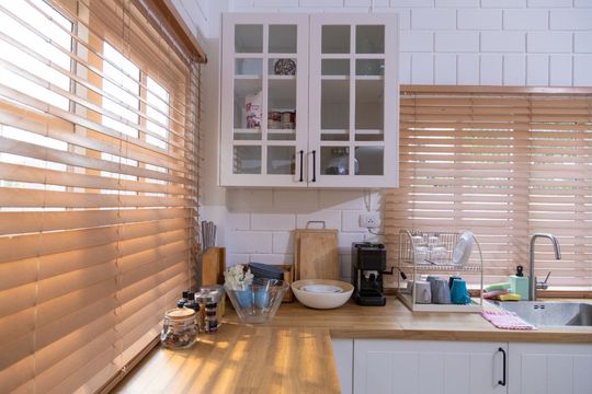 Bright kitchen counter with white cabinets, wooden blinds, sink, coffee maker, and bowls in sunlight