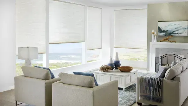 Living room with neutral tones, large windows with shades, and white furniture.