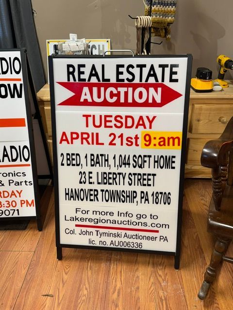 Real estate auction sign with red and yellow text on a wood floor, advertising a Tuesday April 21st 9am sale