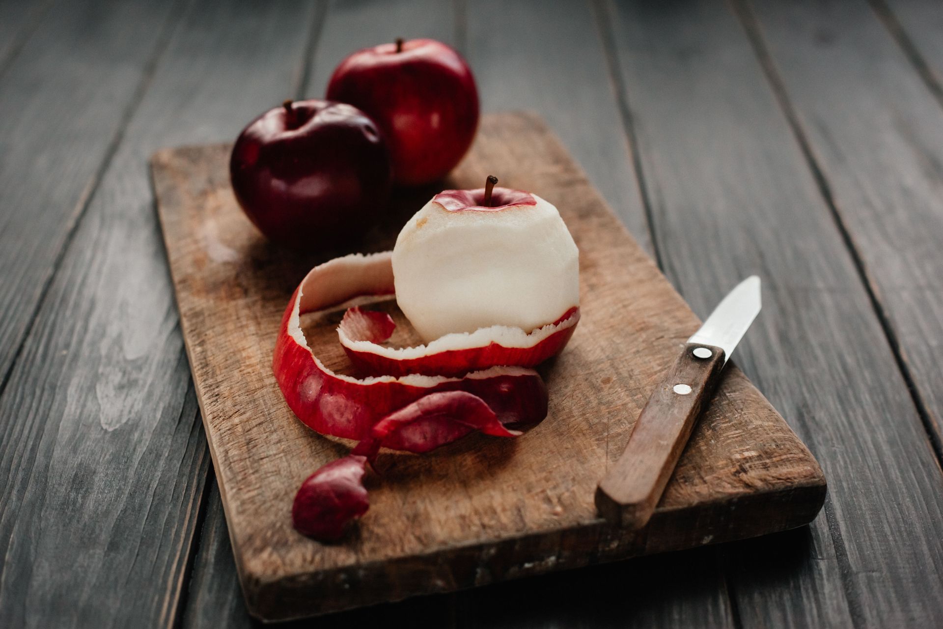 Peeled red apple on a wooden cutting board with a paring knife and two whole apples on a gray table