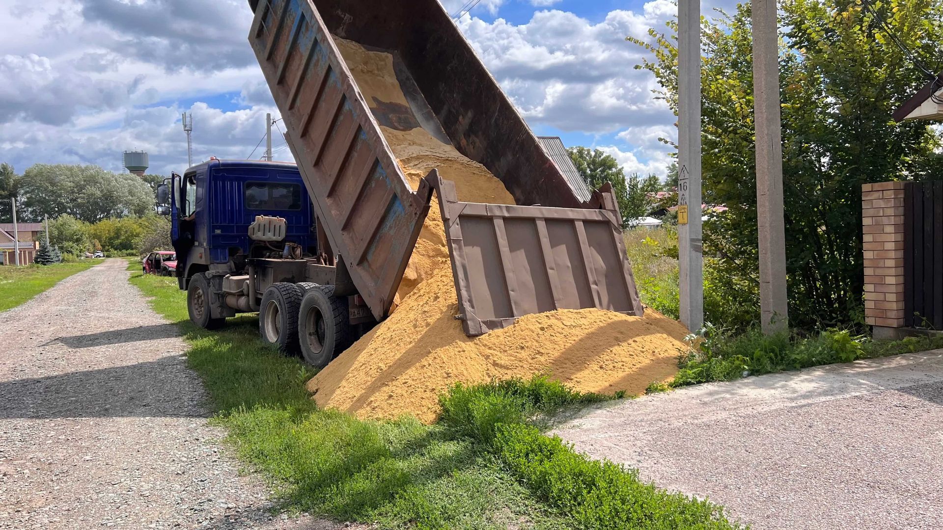 Blue dump truck unloading gravel onto a roadside pile under a partly cloudy sky