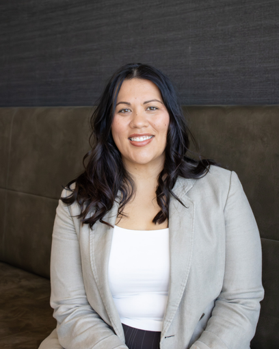 Smiling woman in a light gray blazer and white top, seated against a dark background.