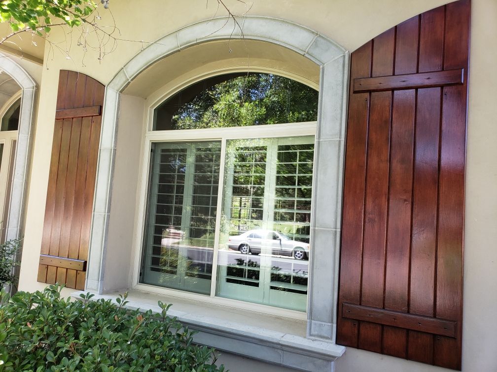 Window with arched top, green door reflection, brown shutters, light stucco exterior, foliage below.