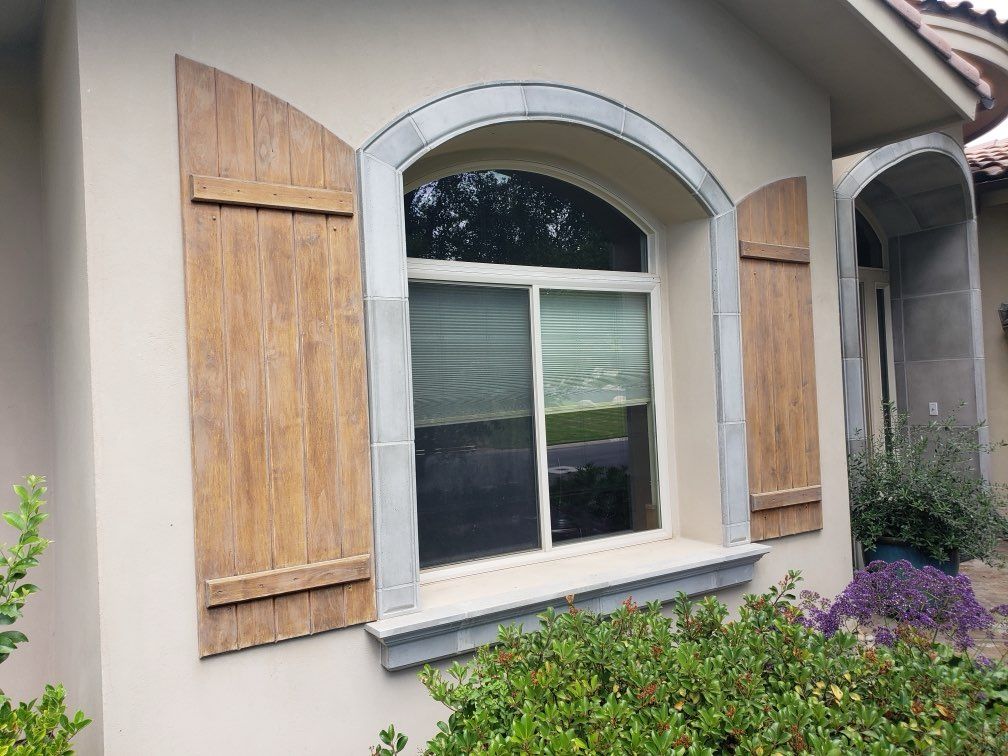 Window with arched top and wooden shutters, light-colored stucco exterior with stone accents and landscaping.