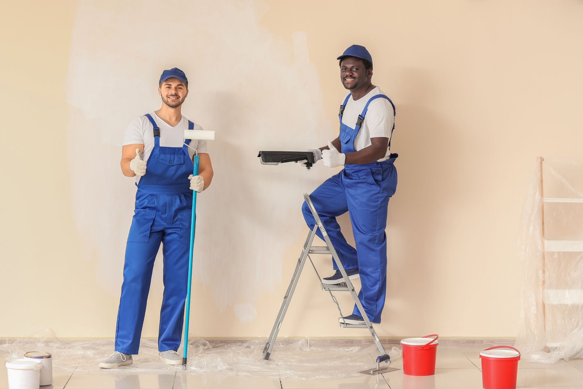 Two painters in blue overalls, one using a roller, the other on a ladder, painting a wall.