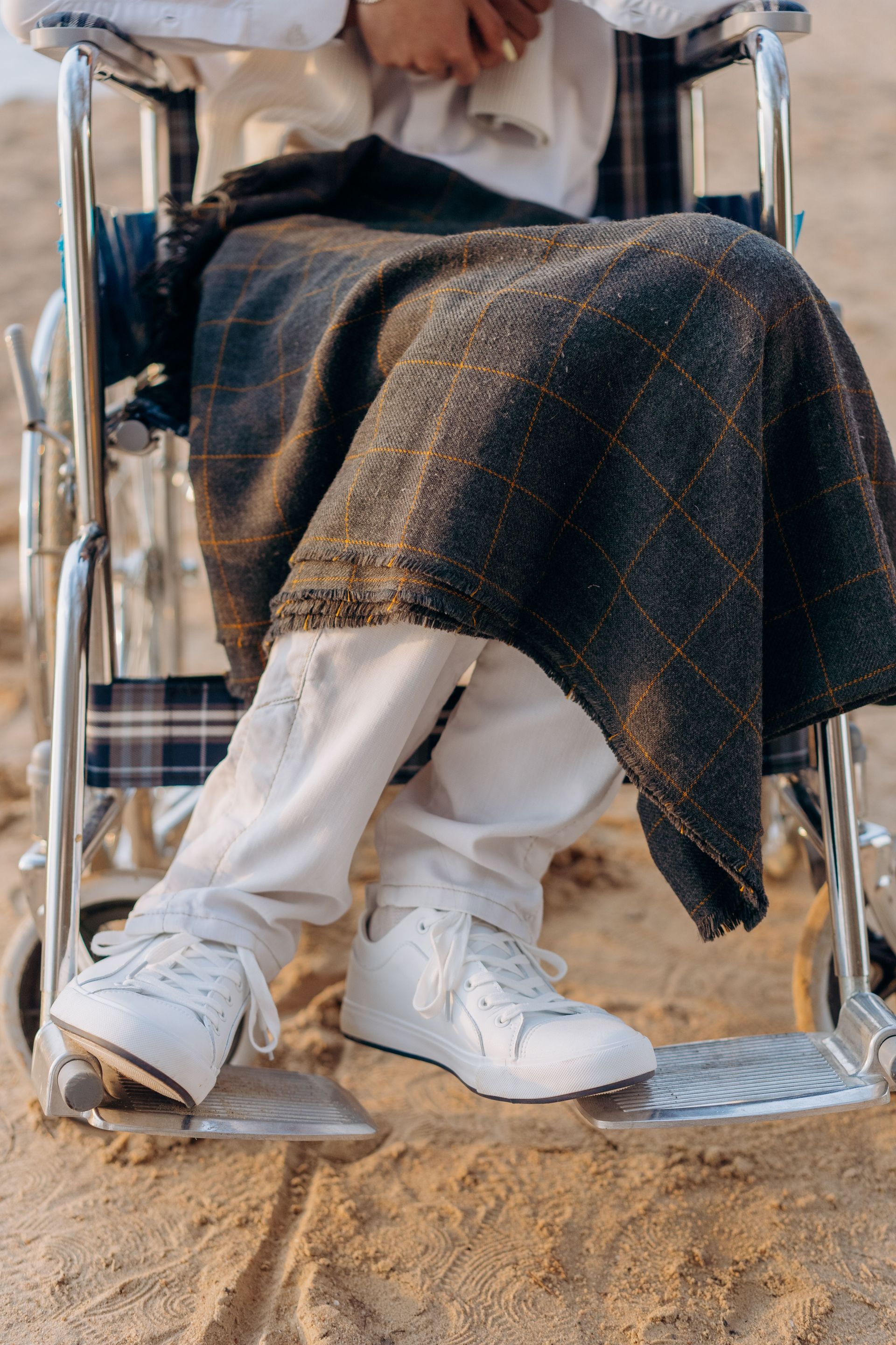 Persoon zit in een rolstoel, draagt witte laarzen en een donkere deken en bevindt zich buiten op een zanderige ondergrond.