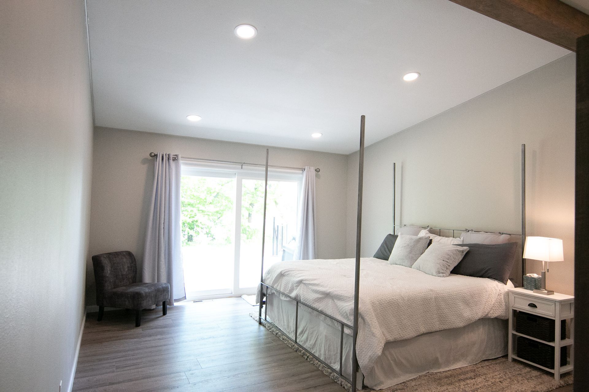 A bedroom with a four poster bed , chair , nightstand and sliding glass doors.