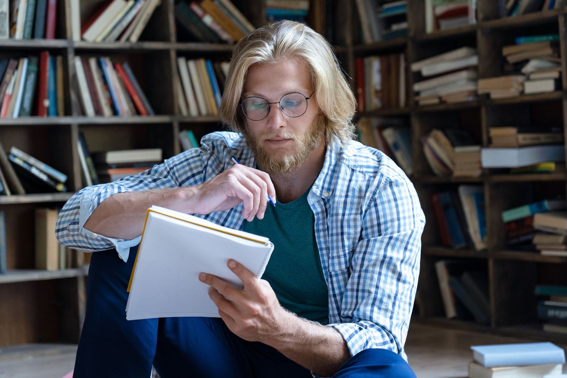 Man with beard and glasses sits on floor, writing in notebook in a library.