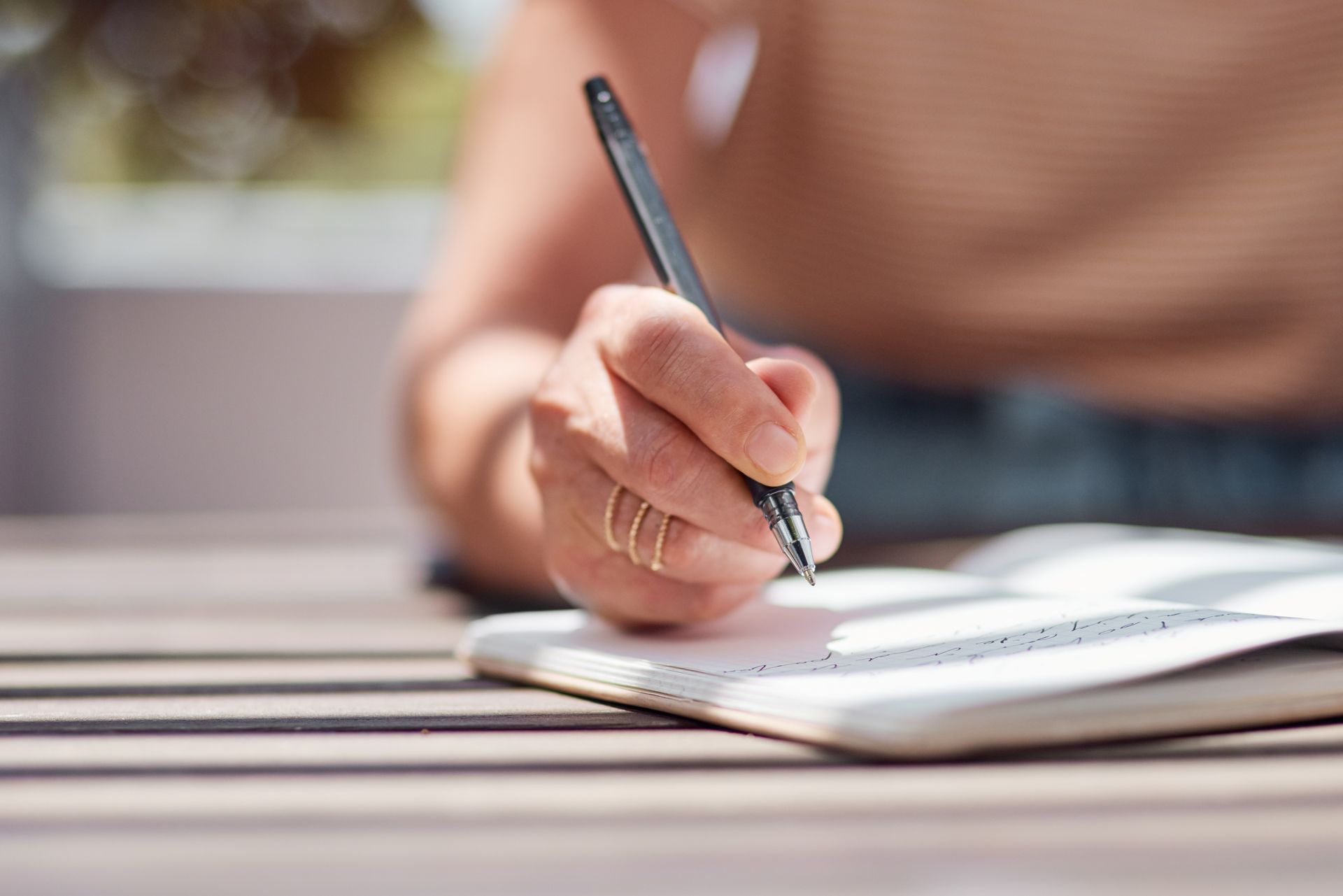 Hand writing in notebook with black pen on a wooden table, outside, sunlight.