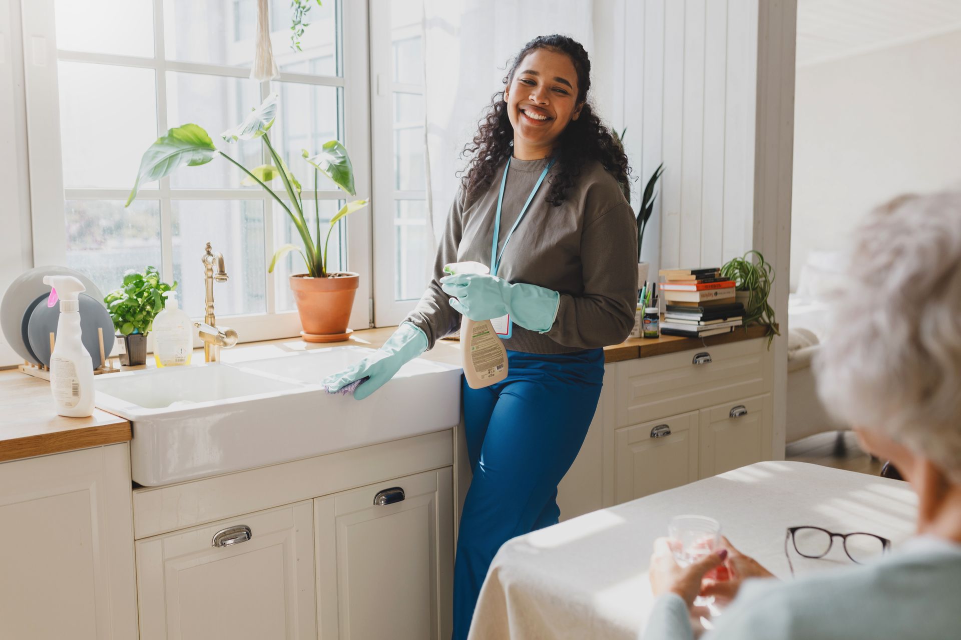 Woman cleaning a kitchen sink, smiling. Wearing gloves and scrubs. Older person sits at a table. Bright room.