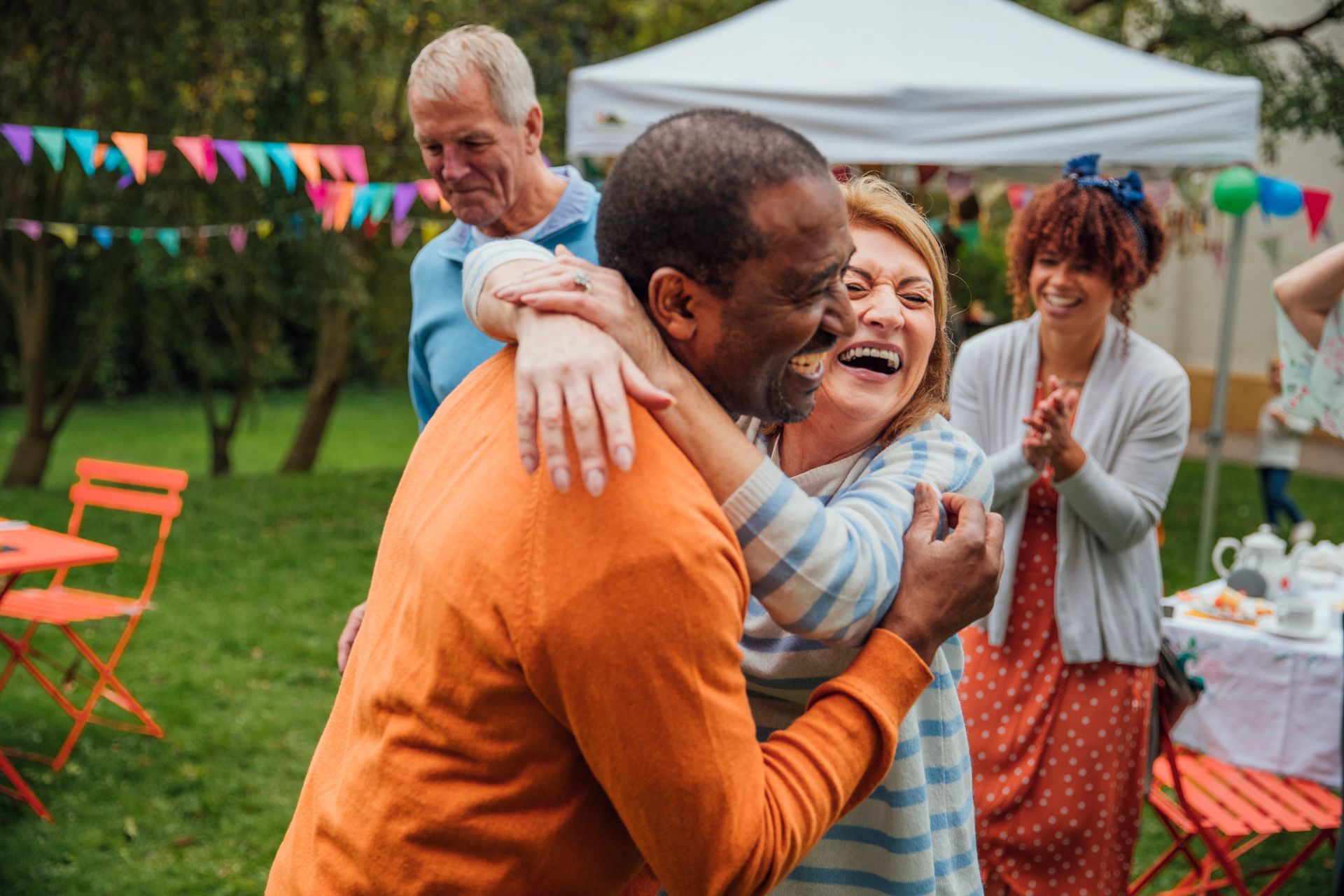 People embracing at an outdoor gathering, smiling. Orange, blue, and white clothing. Festive decorations, sunny day.