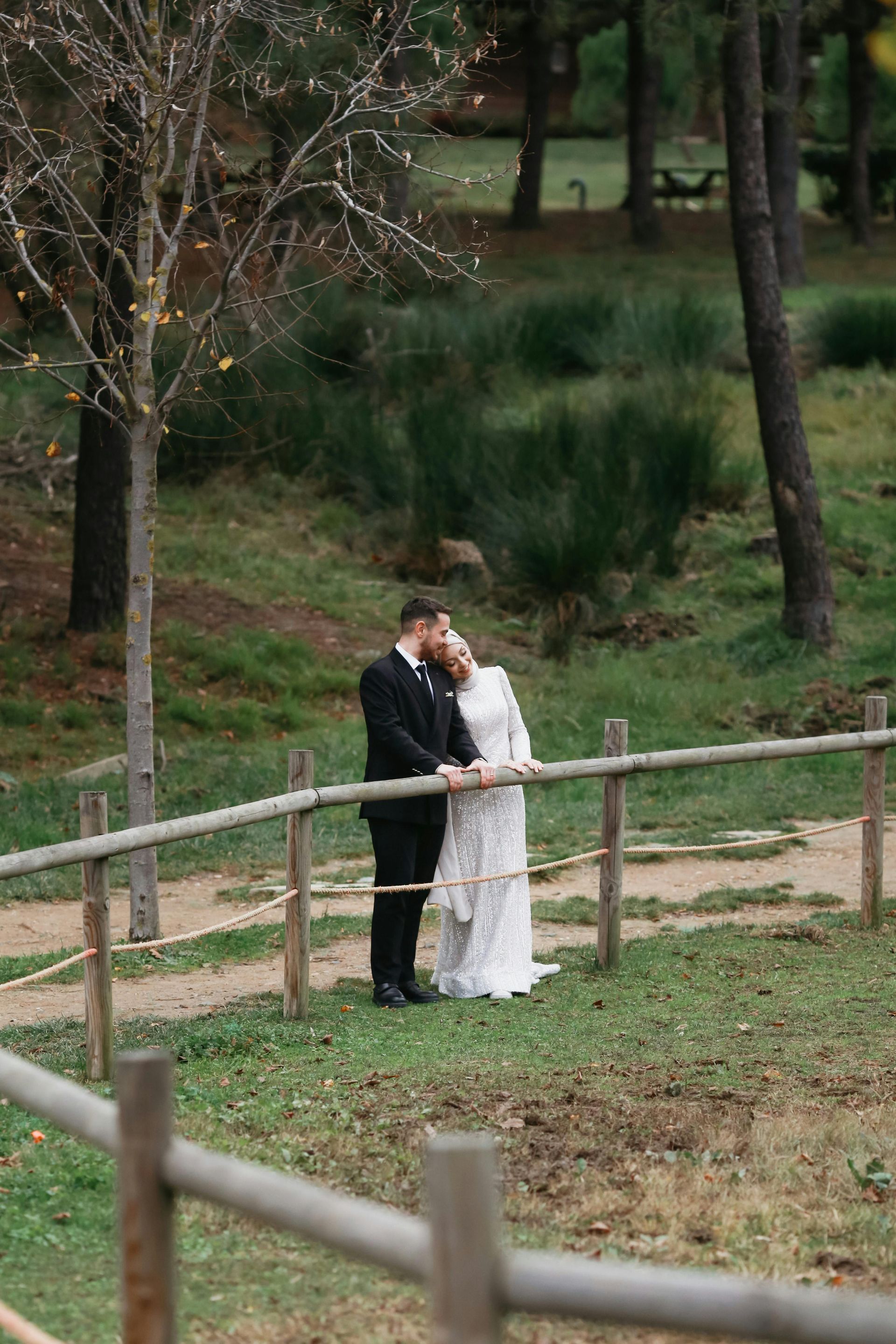 Couple in wedding attire stand near a wooden fence, embracing in a park setting.