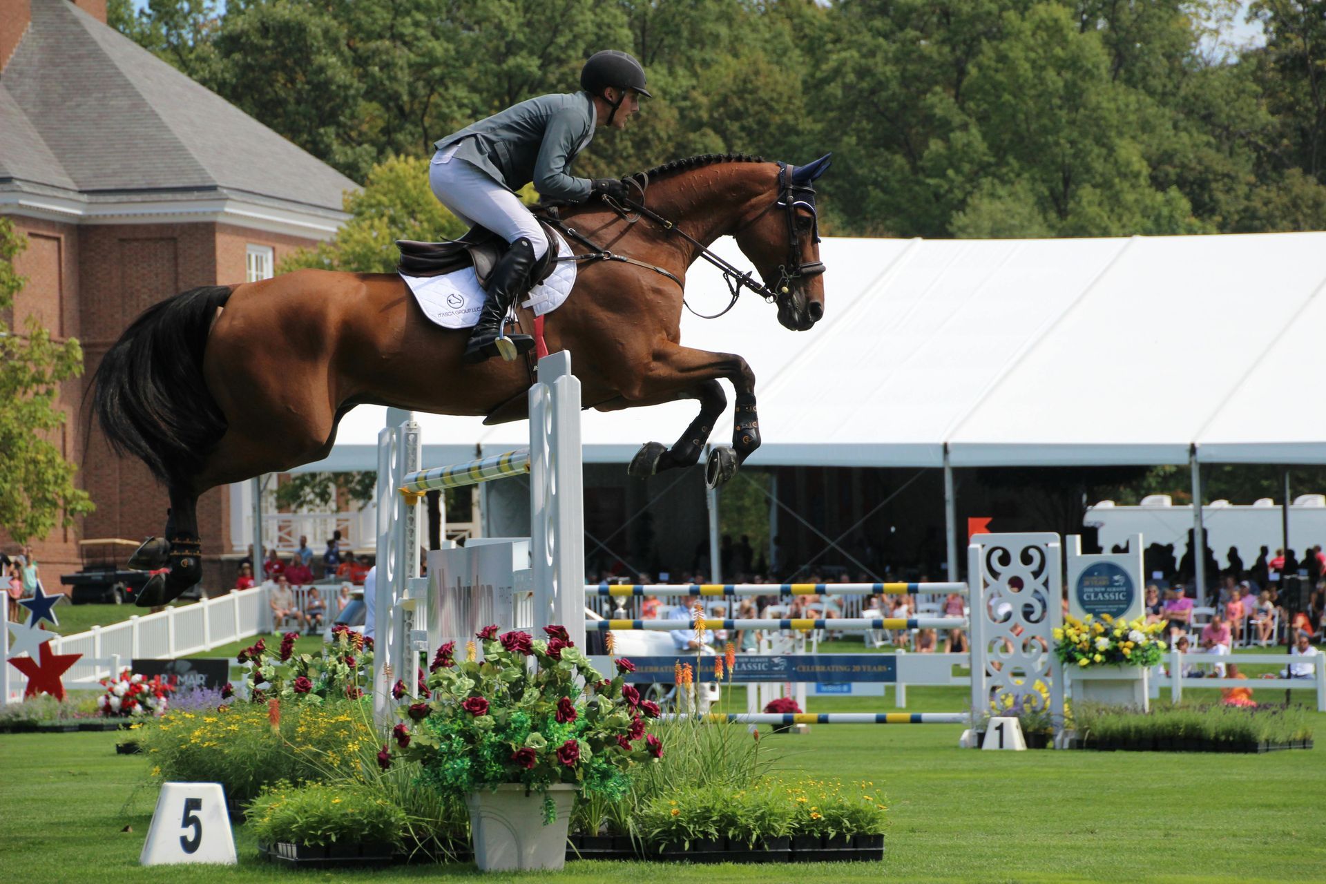 A rider on a brown horse jumps a white fence at an equestrian event with a tent in the background.