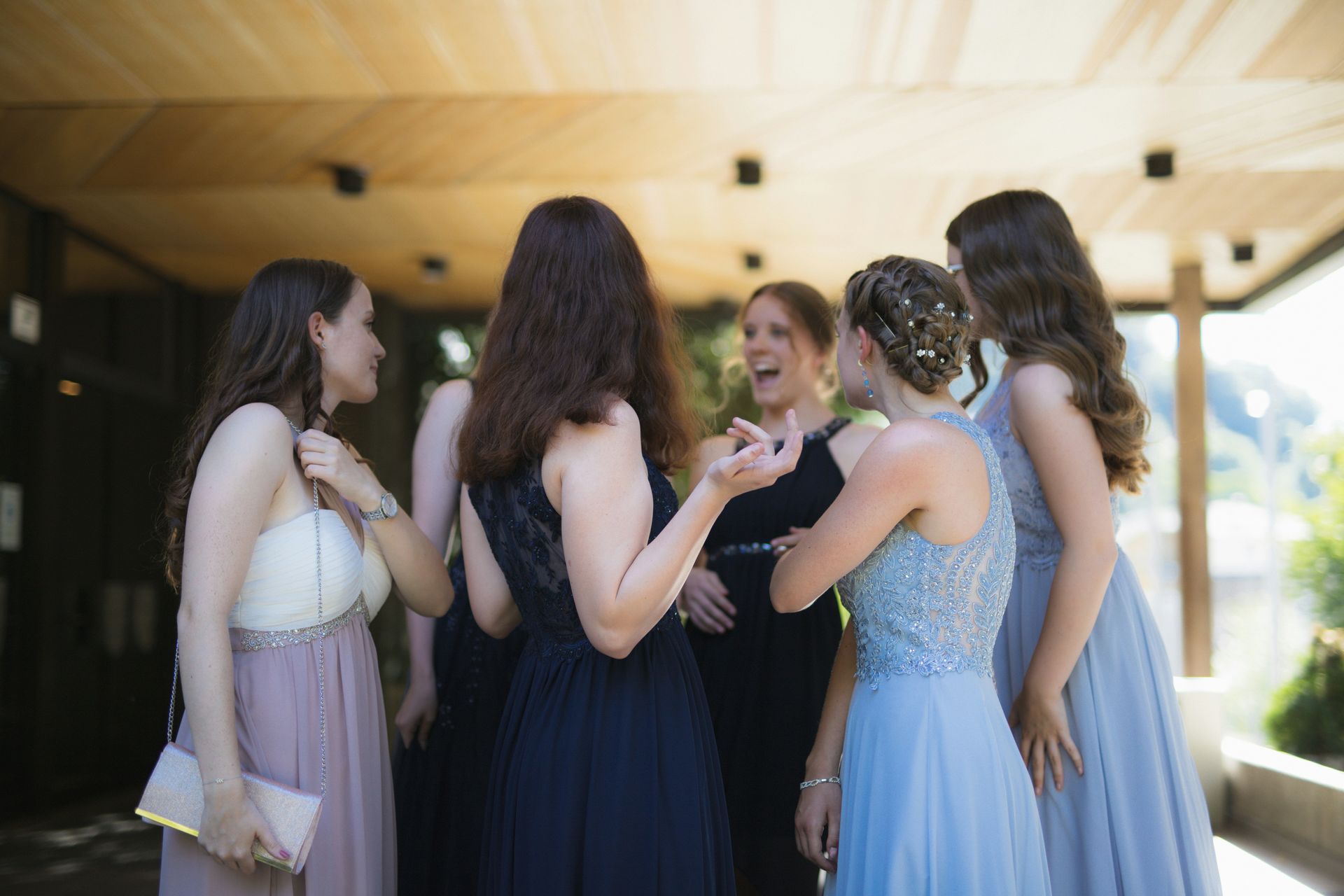 Five women in formal dresses chatting and laughing outdoors under a wooden overhang.