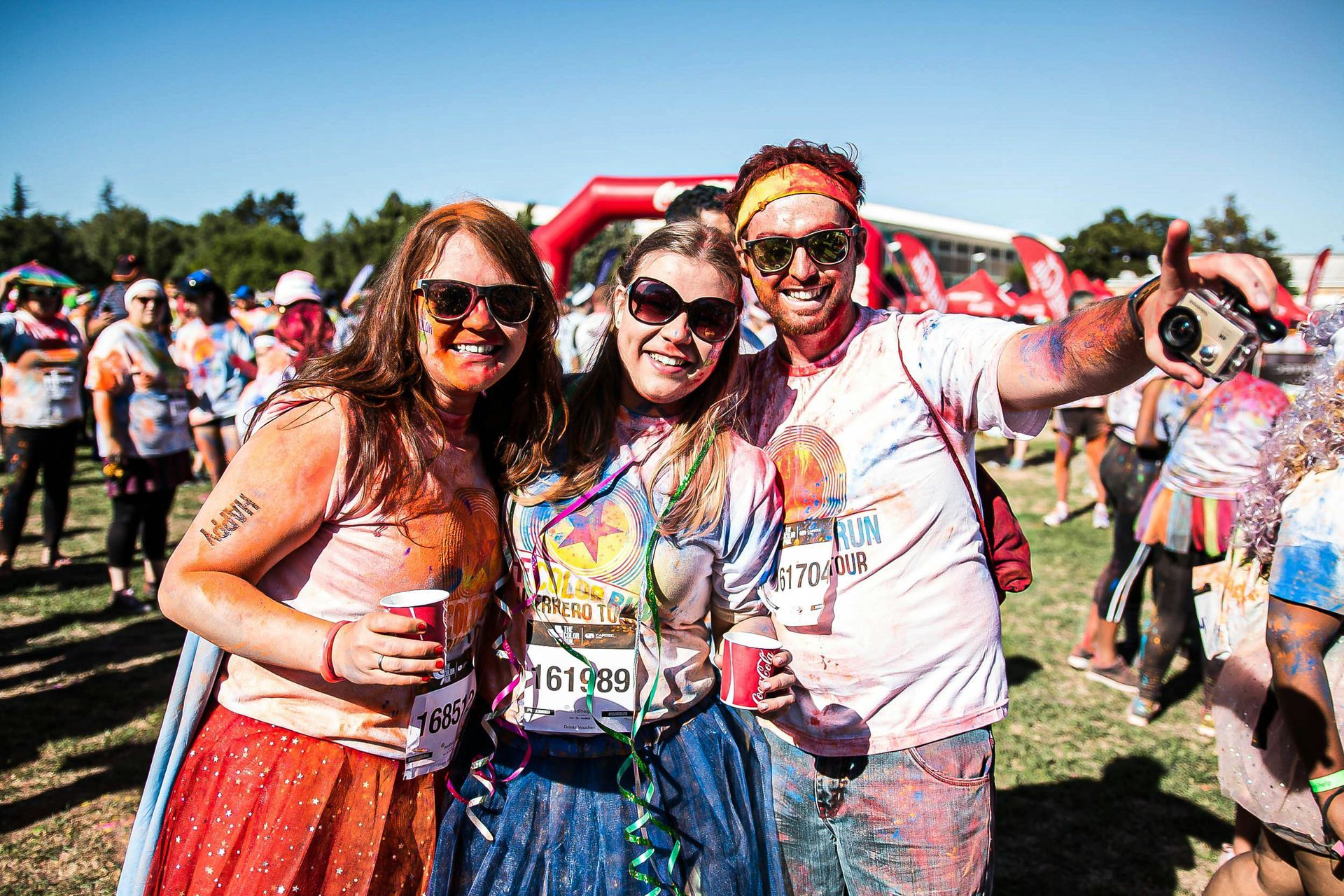 Three people covered in colored powder smile at a color run; outdoors.