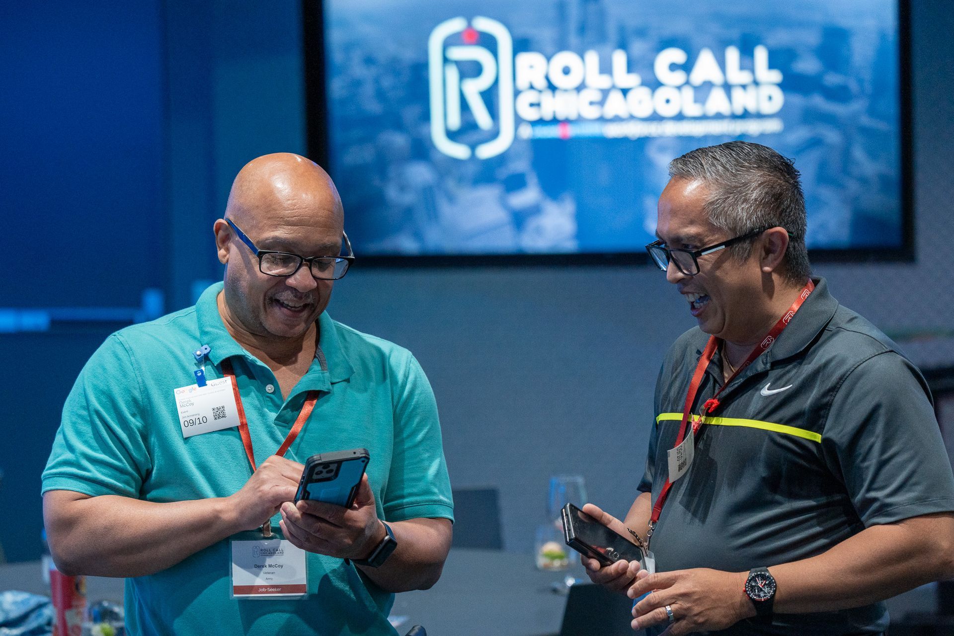 Two men, both wearing name badges, engage in a friendly conversation while looking at their phones.