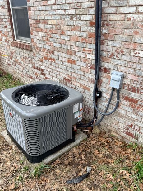 Outdoor air conditioner unit beside a brick house wall with visible wiring and fallen leaves.