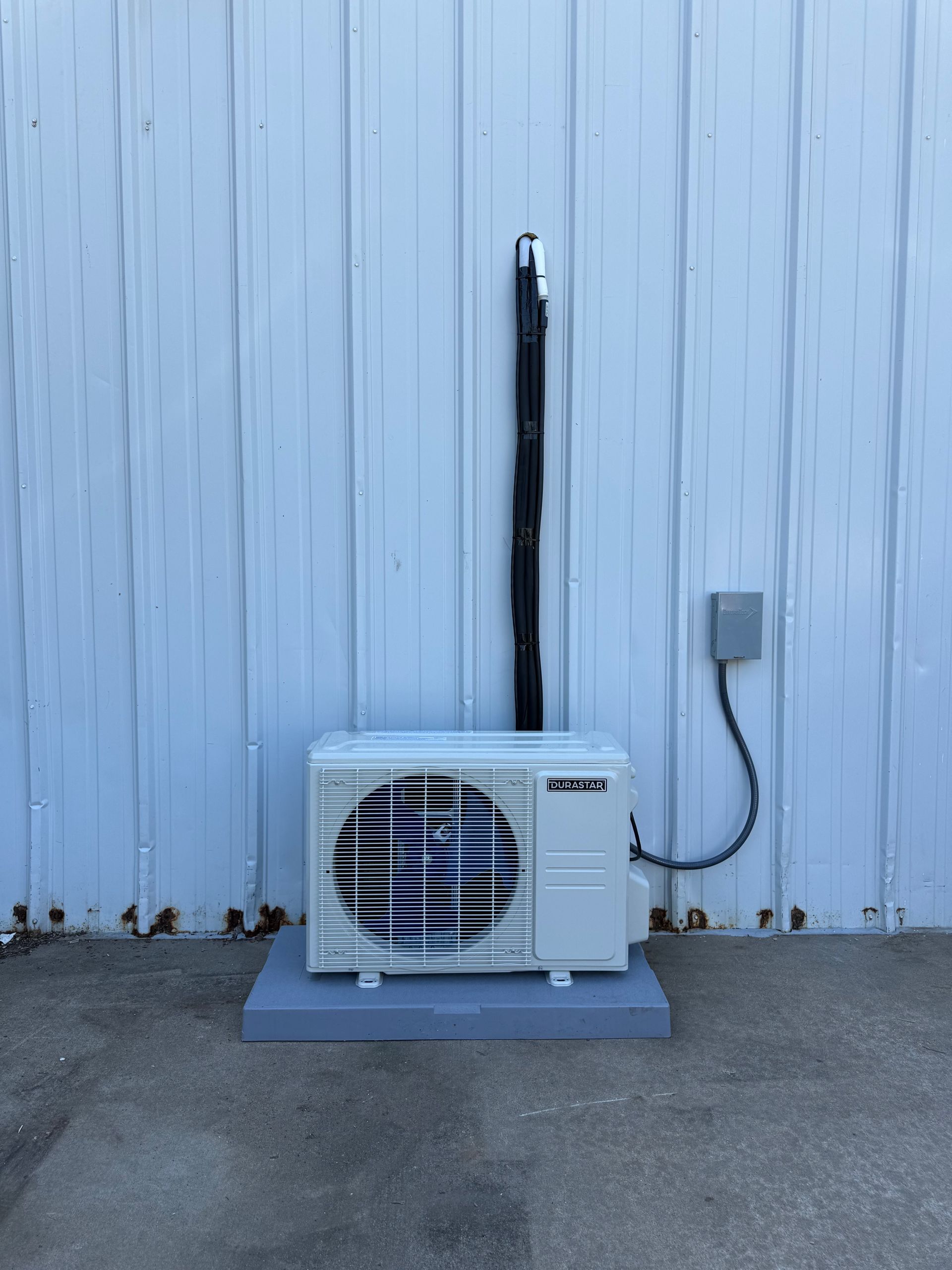 Outdoor HVAC unit on a gray pad against a light blue wall with exposed black piping.