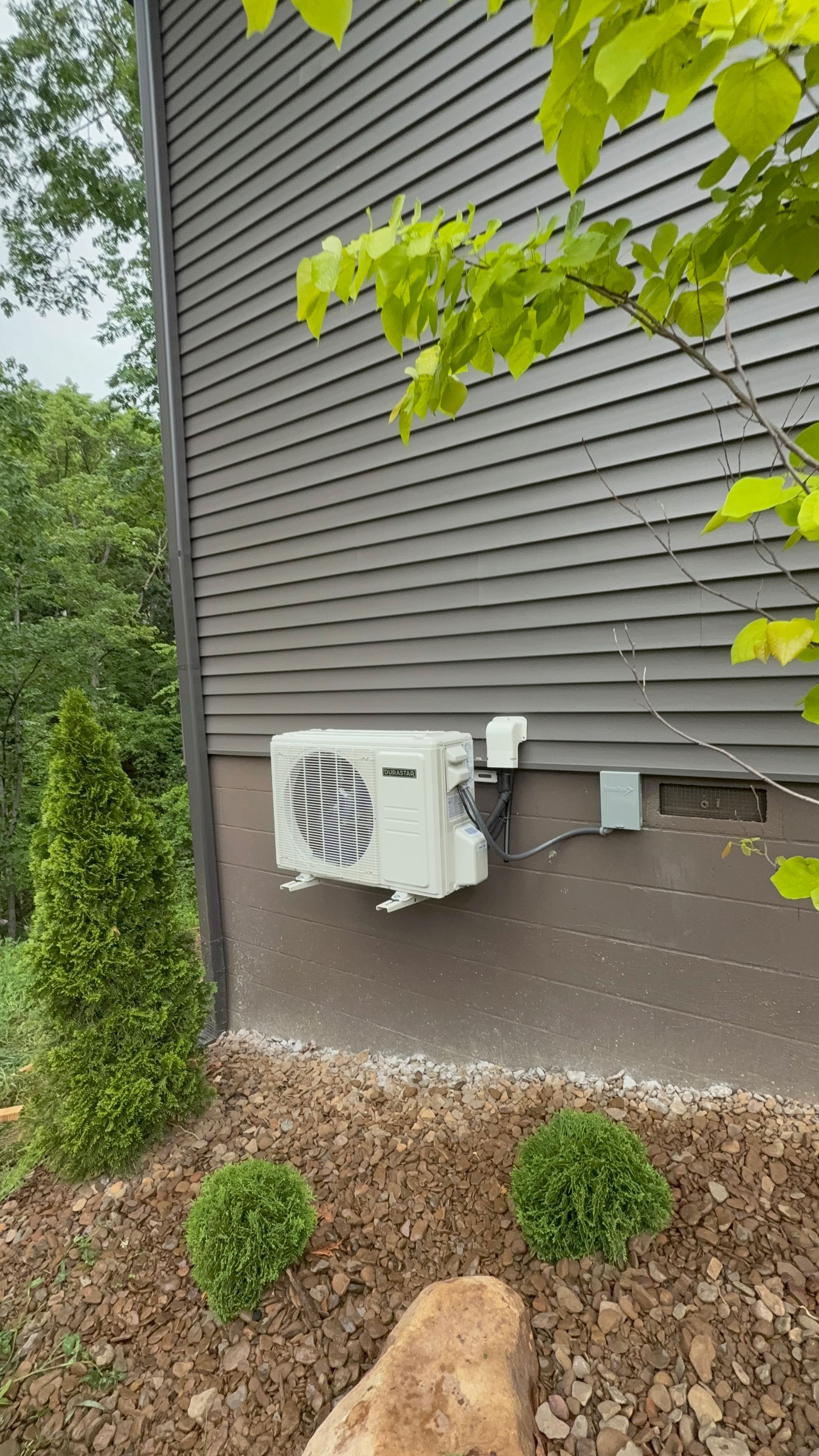 Outdoor wall-mounted HVAC unit beside gray house siding and landscaping shrubs