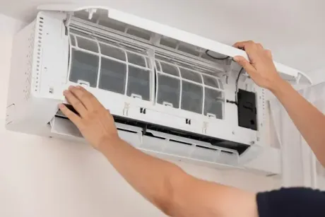 Hands cleaning the open front panel of a wall-mounted air conditioner.