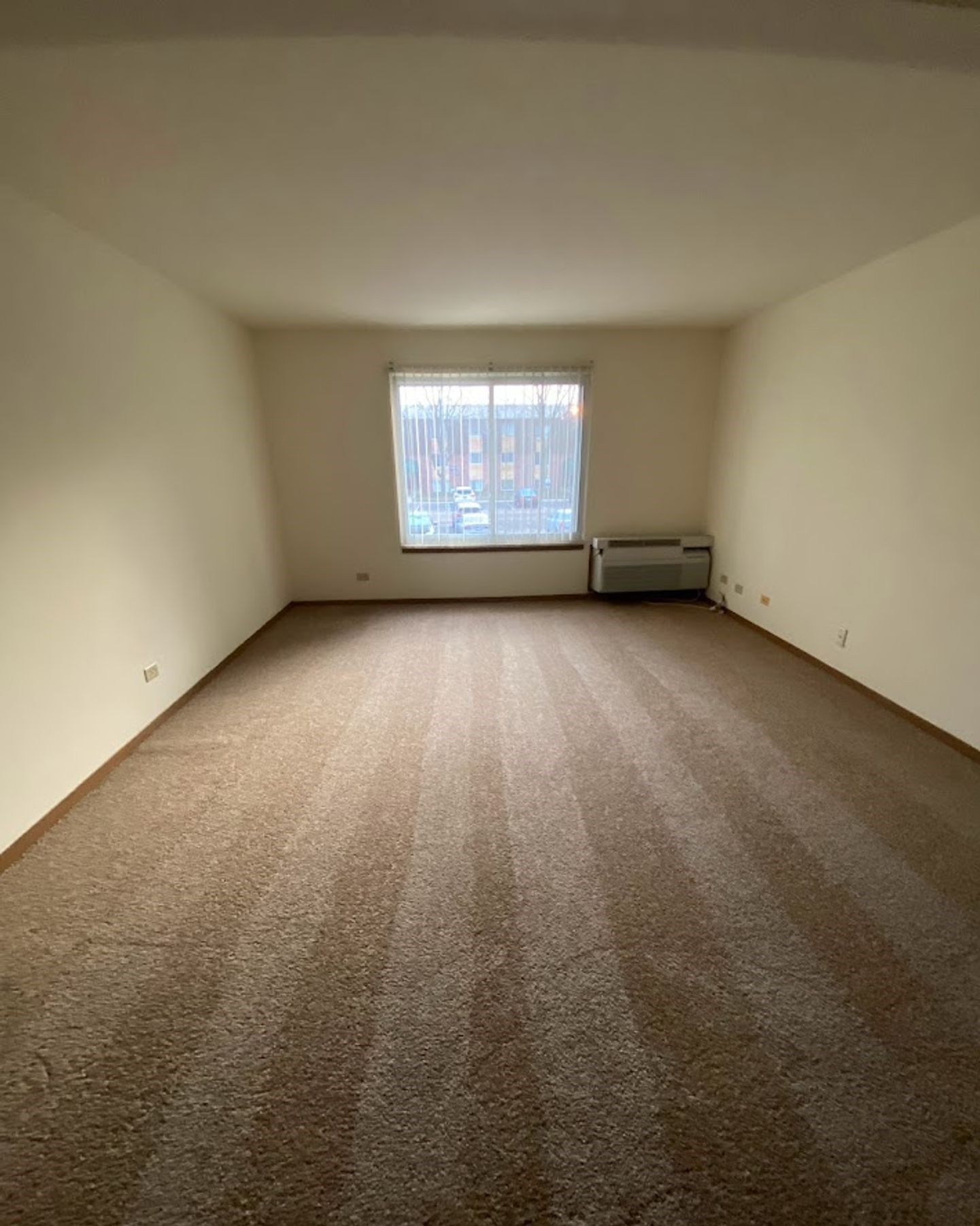 An empty living room with a large window and brown carpet.