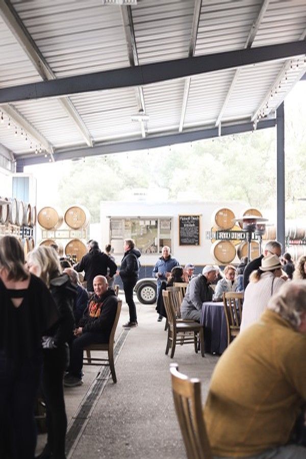 People socialize in an open-air winery patio featuring wooden barrels, a food truck, and tables under a metal roof.