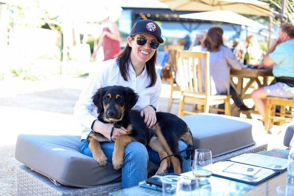 A person wearing sunglasses and a cap sits on an outdoor patio bench, holding a black and tan dog on their lap.