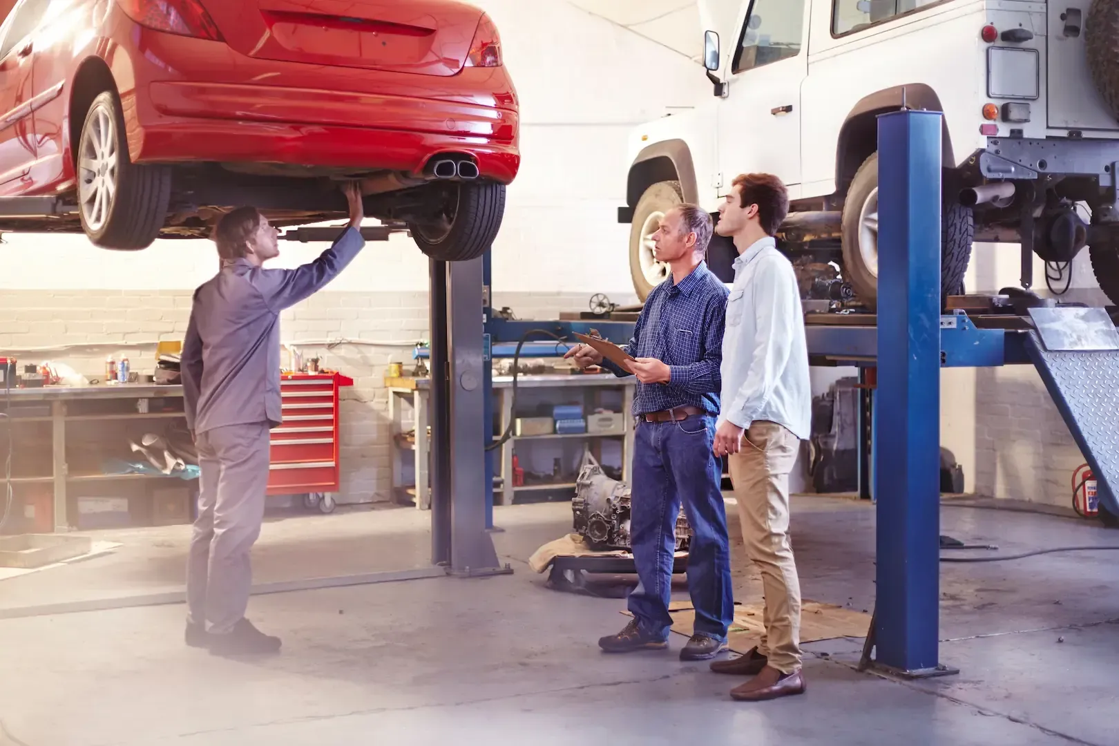 Car raised on a lift in a garage; a mechanic points to the underside as two men watch.