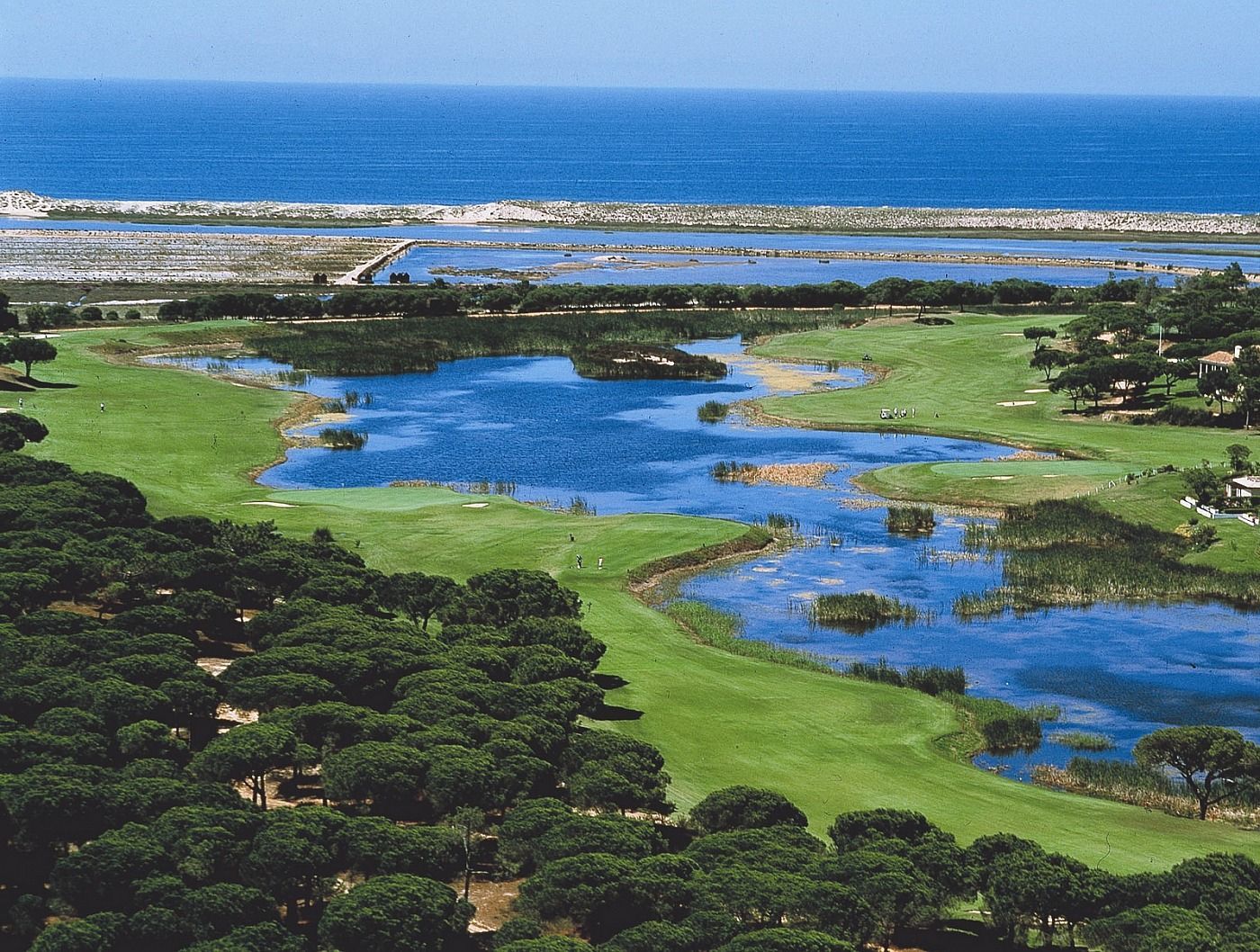 Panoramic view of a lush green golf course fairway under a clear sky, featuring a red flag on a manicured green and strategic sand bunkers.